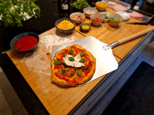 Profi Pizzaschieber mit großer Aluminium-Schaufel und langem Holzgriff beim Herausnehmen einer frisch gebackenen Pizza aus dem Ofen.